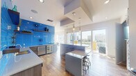 This stylish, single-floor space combines a fully equipped kitchen and an open living area into a cohesive, modern layout. The kitchen is a standout, featuring a bold cobalt-blue subway tile backsplash that extends from counter to ceiling, creating a striking focal point. Warm wood-tone cabinetry with sleek hardware complements white quartz countertops, while stainless steel appliances — including a built-in wine cooler — add a polished, upscale touch. A central island with pendant lighting and bar seating serves as both a functional workspace and a social hub. Warm hardwood flooring flows seamlessly from the kitchen into the living area, unifying the space. The living room is bathed in abundant natural light from expansive floor-to-ceiling glass doors that open to a private balcony overlooking an urban cityscape. A decorative lattice pendant light anchors the seating area, which is designed for relaxed gatherings. The transition between the kitchen and living areas is open and fluid, enhancing the sense of space. A restroom with textured tile accents and recessed lighting is also accessible. Throughout, the design balances vibrant color with clean, minimalist architecture for a bright and inviting atmosphere.