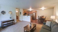 This single-floor space features an open-concept design that seamlessly connects the living, dining, and kitchen areas into one cohesive layout. The living area is finished with plush carpeting and is furnished with a sofa and a wooden coffee table, creating a comfortable and inviting atmosphere. Flush-mounted ceiling light fixtures provide consistent overhead illumination throughout.

The kitchen is positioned centrally and defined by a raised breakfast bar with seating overhang, offering a natural divide between cooking and living spaces. Honey-toned wood cabinetry lines the upper and lower walls, complemented by granite-look countertops. Stainless steel appliances, including a full-sized refrigerator and microwave, add a modern touch. The kitchen area transitions to hardwood-style flooring, distinguishing it from the carpeted zones.

Adjacent to the kitchen, the dining area accommodates a full-sized table and chair set, ideal for everyday meals. A hallway visible in the background suggests access to additional rooms, including what appears to be a bathroom. The overall ambiance is warm, practical, and well-suited for comfortable daily living.