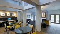 Located in Sterling, Virginia, this single-floor space offers a thoughtfully designed open-concept layout that seamlessly blends functionality with modern style. The interior is characterized by soaring ceilings with angled architectural details and structural columns that define each zone without sacrificing the open feel of the space. The dining area features a sleek round glass-top table surrounded by bold yellow upholstered chairs, providing a striking contrast against the neutral gray and white color palette. Adjacent to the dining space, a well-appointed lounge area includes a comfortable gray sectional sofa, wall-mounted flat-screen television, and built-in dark cabinetry that adds both storage and visual depth. On the opposite side, a cozy seating nook is anchored by a fireplace set against a textured accent wall, with curated artwork adding warmth and character. Recessed lighting and natural light from upper-level windows illuminate the space evenly, enhancing the contemporary ambiance. The patterned carpeting throughout ties the various zones together cohesively, while the overall design strikes a balance between inviting comfort and polished, modern sophistication.