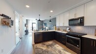This single-floor residence showcases a thoughtfully designed open-concept layout that seamlessly connects the kitchen, dining, and living areas. The kitchen is a standout feature, boasting a two-tone cabinetry scheme with crisp white upper cabinets accented by gold hardware and deep dark lower cabinets, complemented by light quartz countertops and a textured geometric tile backsplash. Black stainless steel appliances, including a gas range, over-the-range microwave, dishwasher, and refrigerator, add a sleek, modern edge. Industrial-style pendant lights hang above the peninsula, which functions as both a prep space and casual dining area. The open living area extends naturally from the kitchen, featuring warm wood-look flooring throughout, a bold accent wall in deep slate gray, and large windows that draw in abundant natural light. The bedroom continues the contemporary aesthetic with a teal accent wall, floor-to-ceiling windows offering outdoor views, and neutral tones that create a calm, restful atmosphere. The bathroom is finished with large-format floor tiles, tall white subway-tiled walls, a glass-enclosed walk-in shower with a built-in bench, and matte black fixtures, delivering a clean and refined look throughout.