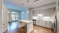 Located at 2925 Buford Dr in Buford, GA, this single-floor residence offers a well-designed, contemporary interior with a cohesive neutral palette throughout. The open-concept kitchen serves as a central highlight, featuring gray shaker-style cabinetry extending to the ceiling, a decorative geometric tile backsplash, quartz countertops, and a full suite of stainless steel appliances including a range, microwave, dishwasher, and refrigerator. A substantial center island with pendant lighting above provides additional workspace and a natural gathering point. The kitchen flows seamlessly into the living area, which is finished with warm hardwood-style flooring, crown molding, and a tray ceiling detail. Large floor-to-ceiling windows and a glass door open to a private balcony, flooding the space with natural light and outdoor views. The bedrooms are carpeted for comfort, with generous proportions and large windows. At least one bedroom features a walk-in closet with built-in shelving. The bathroom is appointed with wood-look tile flooring, a tiled tub-surround, a white soaking tub, and a vanity with under-mount sink and ample cabinetry.
