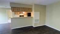 This single-floor space offers a practical and open layout designed to maximize functionality. The living area flows seamlessly toward a pass-through kitchen, connected by a raised countertop bar that serves as both a visual divider and a casual dining or workspace surface. The kitchen is equipped with wood-tone upper cabinetry, a black refrigerator, and a range with an overhead ventilation hood illuminated by a built-in light. The counter space is finished in a light neutral tone, complementing the warm cabinetry above.

The flooring throughout the space features dark hardwood-style planks that provide a rich contrast against the soft, neutral green-toned walls and bright white baseboards. A textured popcorn ceiling adds a classic touch overhead. A large wall-mounted HVAC vent on the main wall ensures climate control throughout the space. The overall color palette is understated and versatile, creating a clean, move-in-ready environment. The open sightlines between the living and kitchen areas contribute to an airy, connected feel well-suited for everyday living.