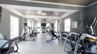 This fitness center offers a comprehensive workout environment designed with both function and aesthetics in mind. The space features a distinctive tray ceiling with a dark wood finish and recessed lighting, lending a polished, upscale feel that sets it apart from a typical gym setting. A central ceiling fixture provides soft ambient light, complemented by natural light streaming in through multiple windows along the walls.

Floor-to-ceiling mirrors line the walls, visually expanding the space and allowing users to monitor their form during exercise. The smooth, light grey flooring provides a clean, durable surface suited for high-traffic fitness use. The neutral grey and white color palette throughout creates a cohesive, modern atmosphere.

The equipment layout is thoughtfully organized. Cardio machines, including ellipticals and treadmills, are arranged along one side, while the central area accommodates an adjustable weight bench and a cable machine for strength training. A well-stocked dumbbell rack along the far wall offers a range of free weights for varied routines. The open floor plan ensures comfortable circulation between stations, making the space practical and accessible for a full-body workout.