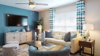 This single-floor residence showcases a well-curated interior design that balances bold color choices with comfortable, functional living spaces. The living room serves as the heart of the home, highlighted by a striking teal accent wall that anchors the entertainment area. Light wood-style flooring flows throughout, while a ceiling fan with integrated lighting adds both practicality and style. Large windows dressed with teal-patterned curtains and horizontal blinds allow generous natural light to fill the room, complementing the neutral tones of the seating arrangement. A textured area rug and dark wood coffee table complete the inviting atmosphere. The kitchen features a two-tone cabinet design — white upper cabinets paired with warm gray lower cabinets — set against a deep navy accent wall. Granite-style countertops and stainless steel appliances, including a dishwasher and refrigerator, give the kitchen a polished, modern feel. The dining area offers an intimate setting with a round glass-top table, white upholstered chairs, and a large abstract artwork, all bathed in natural light from a nearby window. The bedroom is a serene retreat, centered around a bold dark accent wall that frames the bed dramatically, accented by coordinating artwork and warm bedside lighting. Wood-style flooring and a plush navy area rug complete the space.