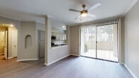 This single-floor residence features a thoughtfully designed open-concept layout connecting the living area and kitchen. The living space is finished with wood-look plank flooring and is anchored by a ceiling fan with a light fixture. Crown molding and a decorative column add architectural detail, while large sliding glass doors fitted with vertical blinds open to a private patio, drawing in natural light and outdoor views.

The kitchen is well-appointed with gray-toned shaker-style cabinetry, light quartz countertops, and a herringbone-pattern tile backsplash. Stainless steel appliances—including a range, microwave, dishwasher, and refrigerator—complement the space, which also features a window above the sink. A pass-through counter connects the kitchen to the living area.

The bedroom offers a ceiling fan, windows with blinds for natural light, and a mix of carpet and wood-look flooring. The bathroom includes a tiled tub-and-shower combination enclosed by a sliding glass door, paired with gray cabinetry for storage. Neutral warm-toned walls throughout the unit create a cohesive and welcoming ambiance.