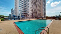 This outdoor amenity space offers a well-appointed pool deck designed for relaxation and recreation in an urban setting. The centerpiece is a generously sized, rectangular swimming pool with crystal-clear turquoise water, accessible via stainless steel handrails and steps. The pool deck is finished in a textured concrete surface that extends broadly in all directions, providing ample room for circulation and leisure. Surrounding the pool, rows of cushioned lounge chairs are neatly arranged beneath large blue umbrellas, offering shade for sunbathers throughout the day. A dedicated dining area features bold red picnic-style tables paired with matching benches and additional oversized umbrellas, creating a comfortable space for outdoor meals and gatherings. Lush container plantings and decorative foliage add touches of greenery throughout the deck. The space benefits from open sky exposure, flooding the area with natural light, while the surrounding high-rise towers provide a striking urban backdrop. Views from the deck extend to neighboring architectural landmarks, mature tree canopies, and the broader cityscape. A gazebo structure and additional seating areas round out this well-equipped outdoor retreat, making it an ideal communal space for residents seeking sun, socializing, and leisure.