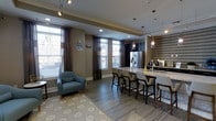 This well-appointed single-floor space showcases a seamless open-concept layout that blends a fully equipped kitchen with a comfortable lounge area. The kitchen is a standout feature, boasting a large island with a sleek granite or quartz countertop and bar-height seating for multiple guests, making it ideal for both casual dining and social gatherings. Dark cabinetry lines the kitchen walls, complemented by a stainless steel French-door refrigerator and elegant pendant lighting that hangs above the island, adding a refined touch to the space. A textured accent wall in a geometric tile pattern provides a striking visual backdrop near the secondary counter area. The lounge zone is anchored by a plush accent chair in a soft blue hue, positioned atop a patterned area rug that adds warmth and texture to the wood-look flooring. Large windows dressed with layered sheer and solid drapes flood the space with natural light while maintaining privacy. Recessed ceiling lighting and track fixtures ensure the space is well-lit at all times. The overall ambiance is modern and inviting, with a neutral warm-gray color palette, cohesive finishes, and thoughtful design details that create a polished, functional environment.