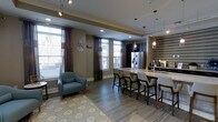 This well-appointed single-floor space showcases a seamless open-concept layout that blends a fully equipped kitchen with a comfortable lounge area. The kitchen is a standout feature, boasting a large island with a sleek granite or quartz countertop and bar-height seating for multiple guests, making it ideal for both casual dining and social gatherings. Dark cabinetry lines the kitchen walls, complemented by a stainless steel French-door refrigerator and elegant pendant lighting that hangs above the island, adding a refined touch to the space. A textured accent wall in a geometric tile pattern provides a striking visual backdrop near the secondary counter area. The lounge zone is anchored by a plush accent chair in a soft blue hue, positioned atop a patterned area rug that adds warmth and texture to the wood-look flooring. Large windows dressed with layered sheer and solid drapes flood the space with natural light while maintaining privacy. Recessed ceiling lighting and track fixtures ensure the space is well-lit at all times. The overall ambiance is modern and inviting, with a neutral warm-gray color palette, cohesive finishes, and thoughtful design details that create a polished, functional environment.