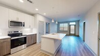 This well-appointed 2-bedroom, 2-bathroom residence offers a clean, modern aesthetic across a single floor of comfortable living space. The open-concept kitchen and living area serves as the heart of the home, featuring light-toned luxury vinyl plank flooring that flows seamlessly throughout. The kitchen is outfitted with stainless steel appliances — including a refrigerator, range, and microwave — complemented by a two-tone cabinet design pairing crisp white upper cabinetry with warm wood-toned lower cabinets. A herringbone tile backsplash adds a refined detail, while a large center island with a white countertop and integrated sink provides both prep space and a casual gathering spot. Pendant lighting with gold-toned fixtures adds a modern finishing touch overhead. The open living area receives natural light through multiple windows with shutters and provides access to an exterior door. Both bedrooms feature plush carpeting and ceiling fans, offering a comfortable retreat. Three closets — including walk-in options with wire shelving — provide generous storage throughout. The bathrooms feature wood-toned vanities with white countertops and wood-look flooring. The overall palette of soft grays, whites, and warm wood tones creates a bright, cohesive, and contemporary atmosphere throughout the residence.