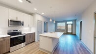 This well-appointed 2-bedroom, 2-bathroom residence offers a clean, modern aesthetic across a single floor of comfortable living space. The open-concept kitchen and living area serves as the heart of the home, featuring light-toned luxury vinyl plank flooring that flows seamlessly throughout. The kitchen is outfitted with stainless steel appliances — including a refrigerator, range, and microwave — complemented by a two-tone cabinet design pairing crisp white upper cabinetry with warm wood-toned lower cabinets. A herringbone tile backsplash adds a refined detail, while a large center island with a white countertop and integrated sink provides both prep space and a casual gathering spot. Pendant lighting with gold-toned fixtures adds a modern finishing touch overhead. The open living area receives natural light through multiple windows with shutters and provides access to an exterior door. Both bedrooms feature plush carpeting and ceiling fans, offering a comfortable retreat. Three closets — including walk-in options with wire shelving — provide generous storage throughout. The bathrooms feature wood-toned vanities with white countertops and wood-look flooring. The overall palette of soft grays, whites, and warm wood tones creates a bright, cohesive, and contemporary atmosphere throughout the residence.