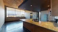 This single-story loft at The Edge showcases a striking industrial aesthetic rooted in its original architectural character. The open living area commands attention with exposed concrete ceilings, polished concrete floors, and a dramatic wall of multi-pane factory-style windows that flood the space with abundant natural light and outdoor views. A partial dropped ceiling detail over the kitchen adds dimension to the open-concept layout.

The kitchen features warm maple cabinetry reaching to the ceiling, granite countertops, a large island with a professional-style coil faucet and undermount sink, and stainless steel appliances including a full-size refrigerator, electric range, and built-in microwave. The bedroom continues the industrial aesthetic with its own factory windows and polished concrete floors.

The bathroom is thoughtfully appointed with slate tile surrounding a glass-enclosed walk-in shower, a vessel sink atop a wood-toned floating vanity, framed mirrors, and warm sconce lighting. An in-unit washer is also included. A dedicated walk-in closet with wire shelving provides generous storage. The overall ambiance balances raw industrial character with refined, contemporary finishes.