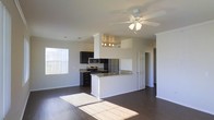This single-story space showcases an open-concept layout that seamlessly connects the living area and kitchen, creating a bright and functional environment. The kitchen is a standout feature, equipped with stainless steel appliances — including a refrigerator, electric range, and microwave — complemented by granite countertops and dark espresso cabinetry. A peninsula counter extends from the kitchen, providing additional workspace and a natural division between the cooking and living areas. Track lighting above the kitchen adds focused illumination to the workspace, while a ceiling fan with integrated lighting in the living area ensures comfortable air circulation and ambient light. The dark hardwood flooring runs consistently throughout the space, offering a rich contrast against the crisp white walls and trim. A large window on the far wall invites generous natural light, casting bright sunlight across the floors and enhancing the open, airy feel of the interior. Crown molding and clean architectural columns add subtle structural character to the space. A paneled door leads to an adjoining room, suggesting additional private spaces beyond the main living area.