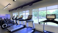 Located at 1000 Cedartree Lane in Claymont, Delaware, this single-floor fitness center offers a well-equipped and inviting space designed for an effective workout experience. The facility is outfitted with a row of modern cardio machines, including treadmills and ellipticals, each equipped with individual digital display consoles for tracking workout metrics. A defining architectural feature of the space is its expansive floor-to-ceiling windows that line the wall, flooding the interior with abundant natural light and providing unobstructed views of the lush, tree-lined greenery outside. The bright and airy atmosphere is further enhanced by roller shades that allow for light control throughout the day. Overhead, a track-style lighting system ensures consistent illumination across the workout floor. The dark, rubberized flooring provides both safety and comfort underfoot, making it well-suited for high-impact exercise. The overall ambiance is clean, modern, and energizing, creating an environment that motivates physical activity while maintaining a connection to the outdoors. This fitness center serves as a thoughtfully designed space that balances functionality with a welcoming aesthetic.