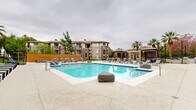 Located at 3100 Van Buren Blvd in Riverside, California, this outdoor amenity space offers a resort-inspired environment designed for relaxation and socializing. At its centerpiece is a generously sized swimming pool with a smooth concrete surround, lined with cushioned lounge chairs and decorative boulder accents — ideal for soaking up the Southern California sun. Adjacent to the pool, a spa is nestled beside a striking blue-accented outdoor fireplace with seating, creating an intimate lounge setting.

The surrounding courtyard is defined by towering palm trees whose canopies frame the space with a tropical ambiance. Strands of bistro lights crisscross overhead, lending a warm, inviting atmosphere for evening gatherings. Modular seating clusters with modern chairs and bold blue accent tables are arranged throughout the paved plaza, encouraging casual conversation and community engagement.

A separate outdoor entertainment area includes BBQ grilling stations and additional shaded seating under pergola-style structures. Manicured green lawns, colorful flower beds, and neatly trimmed hedges line the walkways, adding vibrant natural color throughout. Iron fencing and gated entries provide definition and security without disrupting the open, airy feel of the grounds. The overall design blends functional amenities with a carefully landscaped aesthetic befitting the warm Inland Empire climate.