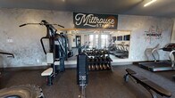 The Millhouse Station fitness center is a thoughtfully designed, single-floor facility that offers a comprehensive workout experience. The space is organized into distinct functional zones, each outfitted with a robust selection of equipment to accommodate a wide range of fitness routines.

The cardio area features multiple treadmills, elliptical machines, and stationary cycling bikes, all positioned in front of a bank of wall-mounted flat-screen televisions for an engaging workout experience. Large windows along one wall flood this zone with natural light and provide views of the surrounding exterior. A ceiling fan overhead contributes to a comfortable environment during intense sessions.

The strength training section includes a multi-station cable machine, selectorized weight machines for targeted muscle groups, a full dumbbell rack, and a functional training area equipped with medicine balls, resistance tools, and a tire for dynamic exercises. Floor-to-ceiling mirrors amplify the sense of space and allow users to monitor their form.

Throughout the facility, durable rubber flooring provides traction and impact absorption. The design aesthetic blends industrial and modern elements, with textured accent walls, dark ceilings, and clean linear lighting. A glass-door entry connects the separate training zones, contributing to an open, airy flow throughout the space.
