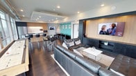 This single-floor space presents a well-designed, open-concept layout that seamlessly integrates multiple functional zones within one expansive area. The living lounge area anchors the space with large, tufted leather sofas arranged around a minimalist white coffee table, all set atop a light area rug contrasting the rich dark hardwood flooring that runs throughout.

A wall-mounted flat-screen television is built into a dark-toned feature wall accented with warm wood paneling, adding a sophisticated focal point to the lounge zone. Recessed ceiling lighting, including a coffered ceiling detail, ensures even, modern illumination across the space.

Toward the rear, a vibrant teal accent wall defines a secondary lounge or meeting area, furnished with additional seating and a flat-screen display, lending a dynamic contrast to the otherwise neutral palette. Adjacent to this, a kitchen or bar area features sleek cabinetry in warm wood tones, a countertop bar with modern black bar stools, and appliances including a microwave, suitable for catering or entertaining. The cohesive design, combining dark tones, wood accents, and contemporary furnishings, creates a polished, professional ambiance throughout.