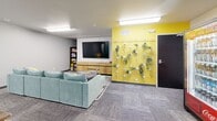 Located at 7175 Dunsmore Rd in Rapid City, South Dakota, this lively communal lounge is a well-appointed gathering space designed for comfort and convenience. The single-floor layout makes efficient use of its compact footprint, offering a variety of zones suited for relaxing and socializing.

The lounge area anchors one side of the room, centered around a plush sectional sofa in a calming seafoam green, paired with a striped area rug and round coffee tables. A wall-mounted flat-screen TV sits above a sleek wooden media console, complemented by a nearby bookshelf stocked with reading materials. On the opposite end, a dining table with dark wooden stools provides a casual space to sit and gather.

One of the room's standout features is its bold, artistic wall treatments. A vibrant yellow accent wall adorned with a vertical living plant display adds a fresh, natural element, while a striking coffee-themed mural painted in teal and brown brings a playful, creative energy to the space.

Practical amenities are thoughtfully integrated, including a beverage vending machine, a snack vending machine, and a dedicated coffee and refreshment station. Overhead fluorescent lighting illuminates the space evenly, and neutral gray carpet tiles throughout tie the room's colorful accents together into a cohesive, welcoming environment.