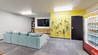 Located at 7175 Dunsmore Rd in Rapid City, South Dakota, this lively communal lounge is a well-appointed gathering space designed for comfort and convenience. The single-floor layout makes efficient use of its compact footprint, offering a variety of zones suited for relaxing and socializing.

The lounge area anchors one side of the room, centered around a plush sectional sofa in a calming seafoam green, paired with a striped area rug and round coffee tables. A wall-mounted flat-screen TV sits above a sleek wooden media console, complemented by a nearby bookshelf stocked with reading materials. On the opposite end, a dining table with dark wooden stools provides a casual space to sit and gather.

One of the room's standout features is its bold, artistic wall treatments. A vibrant yellow accent wall adorned with a vertical living plant display adds a fresh, natural element, while a striking coffee-themed mural painted in teal and brown brings a playful, creative energy to the space.

Practical amenities are thoughtfully integrated, including a beverage vending machine, a snack vending machine, and a dedicated coffee and refreshment station. Overhead fluorescent lighting illuminates the space evenly, and neutral gray carpet tiles throughout tie the room's colorful accents together into a cohesive, welcoming environment.