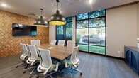 Located at 9310 Otis Ave in Lawrence, Indiana, this conference room is a well-appointed meeting space designed for productivity and style. The room centers around a substantial rectangular wood conference table surrounded by ergonomic modern chairs in white and sage green, comfortably accommodating up to ten guests. Two striking matte black dome pendant lights with gold interiors hang above the table, casting warm, focused light and serving as a bold design statement against the neutral ceiling. A defining feature of the room is the geometric wood mosaic accent wall, which adds rich texture and warmth to the space. Mounted on this wall is a large flat-screen display, making the room well-suited for presentations and collaborative work. Floor-to-ceiling windows along one side of the room flood the interior with abundant natural light and provide open views of the tree-lined surroundings outside. Patterned carpet tiles in muted tones ground the space, while a navy blue credenza and decorative artwork contribute additional character. Recessed lighting supplements the pendant fixtures, and a glass-paneled door connects the room to adjacent areas. The overall ambiance is professional yet inviting, blending contemporary design with functional meeting room essentials.