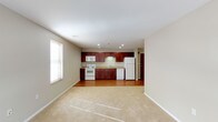This single-floor space offers an open-concept layout that seamlessly connects the kitchen and living area, creating a bright and functional environment. The kitchen is a standout feature, showcasing rich dark-wood cabinetry that provides generous storage alongside a full suite of white appliances, including a refrigerator, electric range, built-in microwave, and dishwasher. A double-basin sink is set beneath under-cabinet lighting, and recessed ceiling lights illuminate the kitchen workspace efficiently. The countertops complement the warm tones of the cabinetry, contributing to a cohesive and inviting aesthetic. The transition from the kitchen flows naturally into the open living area, which is finished with plush, neutral-toned carpeting that adds warmth underfoot. A large window fitted with horizontal blinds allows natural light to fill the space, enhancing the airy feel of the interior. The walls are finished in a clean, neutral tone that reflects light and creates a sense of openness throughout. Smooth white ceilings with recessed lighting throughout provide consistent, well-distributed illumination. The overall ambiance is clean, practical, and welcoming, making this a comfortable and versatile single-floor living space.