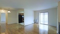 This single-floor space offers a straightforward, functional layout with distinct living and kitchen areas. The living area is generously proportioned, featuring warm hardwood flooring that runs throughout and reflects natural light from the large sliding glass door. This floor-to-ceiling glass door provides direct access to an outdoor balcony or patio, filling the interior with abundant daylight and offering a view of the exterior surroundings. Neutral white walls create a clean, versatile backdrop throughout the space. A wall-mounted air conditioning unit is positioned near the sliding door, providing climate control for the living area. Baseboard heating runs along the perimeter walls, indicating a dual-system approach to year-round comfort. The kitchen is partially open to the living area through a wide doorway, allowing for visual connectivity between the two spaces. It is equipped with warm-toned wood cabinetry, a black refrigerator, and a dark range, offering a functional cooking environment. The overall ambiance of the space is bright and understated, with its hardwood flooring and natural light serving as the primary design highlights.