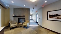 This single-floor space presents a thoughtfully designed lobby or common area that balances comfort and functionality. The centerpiece of the lounge area is a floor-to-ceiling tiled fireplace, clad in sleek, vertically stacked gray tile with a dark wood mantel, creating a warm and inviting focal point. A pair of striped armchairs and a small round side table are arranged in front of the fireplace, offering a comfortable seating arrangement for guests or visitors. The flooring features a patterned carpet that adds texture and a sense of warmth to the space. Recessed lighting is installed throughout the ceiling, providing consistent, clean illumination across the area. A wide corridor extends from the lounge toward a set of glass entry doors, which allow natural light to flow into the space and create an open, airy feel. The walls are finished in a neutral tone, contributing to the bright and professional atmosphere. Security and safety features, including overhead cameras and clearly marked emergency exit signage, are visibly integrated into the design. A wall-mounted display screen is positioned near the seating area, adding a functional element to the space. Overall, the layout and finishes suggest a well-maintained, professional environment suited for a commercial or hospitality setting.