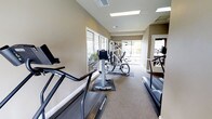 This fitness center offers a clean, functional workout environment designed to support a range of exercise routines. The space is finished with neutral beige walls and carpeted flooring, creating a comfortable and welcoming atmosphere. Overhead, recessed fluorescent lighting is complemented by natural light streaming in through large windows along one wall, providing an open and airy feel throughout. The equipment layout is thoughtfully organized, featuring multiple treadmills positioned for ease of use, a multi-station strength training machine, and additional cardio equipment. The machines are arranged with enough spacing to allow users to move comfortably between stations. A mirrored wall on one side enhances the sense of space and allows for form checks during exercise. Ventilation is supported by ceiling vents, ensuring a consistent and comfortable environment during workouts. The overall design is practical and straightforward, with a focus on functionality and accessibility. This fitness center provides residents or members with a convenient, on-site option for maintaining an active lifestyle without the need to travel to an external facility.