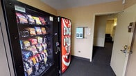 This single-floor space is designed with guest convenience in mind, centered around a well-equipped vending area. The vending alcove features a fully stocked snack machine offering a wide variety of chips, crackers, and packaged treats, alongside a Coca-Cola beverage dispenser providing a selection of cold drinks. The walls are finished in a warm neutral tone, complemented by dark carpeted flooring that runs throughout the corridor. A textured ceiling adds a classic touch to the utilitarian space. A wall-mounted informational panel or key access unit is positioned nearby, suggesting this space serves guests of a hospitality or lodging facility. A doorway leads further into the property, connecting the vending area to additional rooms or corridors beyond. The layout is straightforward and functional, making it easy for guests to navigate. Overall, the space prioritizes practicality and accessibility, ensuring that essential refreshments and services are readily available in a clean, no-frills environment.