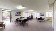 This well-appointed single-floor space is designed to accommodate a variety of gathering and meeting needs. The main area is a generously sized open room featuring patterned carpet flooring and bright overhead fluorescent lighting. Multiple round tables with chairs are arranged throughout, offering flexible seating configurations for group meetings or social events. Large windows fitted with horizontal blinds line the walls, allowing natural light to filter in and provide views of the surrounding exterior. Colorful artwork depicting water scenes adorns the walls, adding a welcoming visual touch.

A compact kitchenette is included, outfitted with wood-tone cabinetry, a range, a sink, a microwave, and ample counter and storage space — ideal for catering or light food preparation. Adjacent to the main room is a cozy lounge area anchored by a traditional wood-mantled fireplace, complemented by wingback chairs, a round table with seating, and a framed landscape painting above the mantle. Glass doors connect this lounge to an outdoor area, further enhancing the sense of openness.

The space also includes an accessible restroom featuring grab bars, a wall-mounted sink, and tile flooring. A brick accent wall visible in the layout adds architectural character. Overall, this space presents a functional and inviting environment suited for meetings, events, or community use.