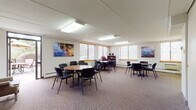 This well-appointed single-floor space is designed to accommodate a variety of gathering and meeting needs. The main area is a generously sized open room featuring patterned carpet flooring and bright overhead fluorescent lighting. Multiple round tables with chairs are arranged throughout, offering flexible seating configurations for group meetings or social events. Large windows fitted with horizontal blinds line the walls, allowing natural light to filter in and provide views of the surrounding exterior. Colorful artwork depicting water scenes adorns the walls, adding a welcoming visual touch.

A compact kitchenette is included, outfitted with wood-tone cabinetry, a range, a sink, a microwave, and ample counter and storage space — ideal for catering or light food preparation. Adjacent to the main room is a cozy lounge area anchored by a traditional wood-mantled fireplace, complemented by wingback chairs, a round table with seating, and a framed landscape painting above the mantle. Glass doors connect this lounge to an outdoor area, further enhancing the sense of openness.

The space also includes an accessible restroom featuring grab bars, a wall-mounted sink, and tile flooring. A brick accent wall visible in the layout adds architectural character. Overall, this space presents a functional and inviting environment suited for meetings, events, or community use.