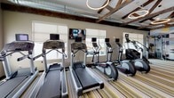 This fitness center combines industrial-chic design with modern amenities to create an inspiring workout environment. The most striking architectural feature is the soaring ceiling adorned with exposed dark timber trusses and metal riveted beams, which lend a bold, warehouse-inspired aesthetic to the space. Suspended circular pendant lights provide warm, focused illumination throughout.

The flooring consists of multi-toned striped carpeting in shades of gold, grey, and white, adding a dynamic visual element underfoot. One wall is dominated by a large, eye-catching geometric mural featuring a mosaic of triangular shapes in muted blues, greys, and yellows, serving as a colorful focal point. Floor-to-ceiling mirrors on adjacent walls enhance the sense of openness and allow users to monitor their form during workouts.

Natural light filters in through multiple windows fitted with roller shades, keeping the atmosphere bright and airy. The cardio zone is well-stocked with treadmills, elliptical trainers, and stationary bikes — all equipped with individual screens. The strength-training area offers a variety of cable machines, selectorized weight stations, free weights, and benches. A dedicated open area with yoga mats provides space for stretching and flexibility work. The combination of thoughtful design and comprehensive equipment makes this a fully functional and visually engaging fitness facility.