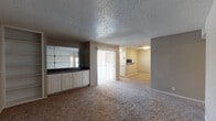 This single-floor unit offers a practical and functional layout spread across several well-defined areas. The living room serves as the central gathering space, featuring wall-to-wall carpet in a neutral tone and a textured popcorn ceiling. A standout built-in unit lines one wall, combining white cabinetry with a dark countertop and mirrored, glass-shelved display sections above — adding visual depth to the room. A sliding glass door with vertical blinds provides access to the outdoors and brings in natural light. The layout connects seamlessly to a separate carpeted room, visible through an open passage, which functions as an additional living or dining area with a ceiling-mounted light fixture.

The galley-style kitchen is efficiently arranged with white cabinetry, granite-look countertops, wood-look plank flooring, and black appliances including a range, microwave, refrigerator, and dishwasher. The bathroom features a wide vanity with white cabinetry, a granite-look countertop, a large mirror, a soaking tub with shower, and wood-look tile flooring. The bedrooms are carpeted with neutral walls; one includes a ceiling fan with light fixture and a window for natural light, while another offers a bright corner window. Throughout, the unit maintains a consistent, neutral color palette.