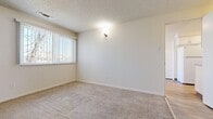 This single-floor unit offers a practical and functional layout across four rooms, finished in a clean, neutral palette throughout. The main living area features plush wall-to-wall carpeting, crisp white walls, and a textured ceiling, creating a cozy and understated atmosphere. A wall-mounted light fixture adds a subtle accent, while vertical blinds on the window allow for adjustable natural light and outdoor views. The galley-style kitchen is efficiently designed, featuring white upper and lower cabinetry, light granite-style countertops, a white range, and a full-size refrigerator. Wood-look vinyl flooring runs the length of the kitchen, providing a durable and easy-to-maintain surface. A secondary door in the kitchen provides additional access to the unit. An additional room, visible through a doorway, includes a compact refrigerator and tiled flooring, suggesting use as a utility or supplementary space. The entry area is defined by a solid white door with double knob hardware. Throughout the unit, consistent white trim, baseboards, and neutral wall tones contribute to a cohesive and tidy appearance. The overall layout is straightforward and well-suited for efficient everyday living.