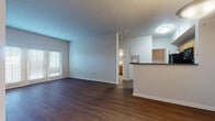 This single-floor residence offers a well-organized layout that balances open communal areas with private living spaces. The living area is expansive and airy, finished with rich, dark hardwood-style flooring that extends throughout the main areas. Natural light filters in through blinded windows and a set of French doors, lending the space a bright and welcoming atmosphere. A half-wall peninsula separates the living area from the kitchen, maintaining an open, connected feel.

The kitchen is well-appointed with warm honey-toned wood cabinetry offering generous storage, dark granite-style countertops, and a full suite of black appliances including a refrigerator, electric range, and over-the-range microwave. Light wood-style flooring completes the kitchen's practical aesthetic.

A hallway connects the main living areas to the private spaces, transitioning from hardwood-style flooring to tile as it approaches a laundry area. The bedrooms feature plush wall-to-wall carpeting and windows with blinds that allow natural light while maintaining privacy.

The bathroom is clean and functional, featuring a wood-toned vanity with an integrated sink, a large mirror, and a full tub and shower combination enclosed by a curtain with tiled surround. The neutral gray palette throughout maintains a cohesive, modern tone.