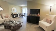 This single-floor residence offers a thoughtfully designed open-concept layout that seamlessly connects the living and dining areas, creating a spacious and cohesive environment ideal for everyday living and entertaining. The living area features soft, plush wall-to-wall carpeting that adds warmth and comfort underfoot, complemented by neutral-toned walls that create a bright and inviting atmosphere. A media console serves as a focal point, providing ample storage and display space. The adjacent dining area is positioned to take full advantage of a large sliding glass door or wide window, allowing generous natural light to pour into the space and offering views to the outdoors. A decorative round mirror and coordinating dark-wood furniture pieces add a touch of modern elegance throughout. The cohesive color palette of warm neutrals, accented by bold pops of orange décor, ties the spaces together harmoniously. The open flow between rooms enhances the overall sense of space and scale, making the layout feel expansive and functional. Ceiling-mounted lighting provides even illumination throughout, ensuring the space remains bright and welcoming at any time of day.