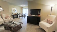 This single-floor residence offers a thoughtfully designed open-concept layout that seamlessly connects the living and dining areas, creating a spacious and cohesive environment ideal for everyday living and entertaining. The living area features soft, plush wall-to-wall carpeting that adds warmth and comfort underfoot, complemented by neutral-toned walls that create a bright and inviting atmosphere. A media console serves as a focal point, providing ample storage and display space. The adjacent dining area is positioned to take full advantage of a large sliding glass door or wide window, allowing generous natural light to pour into the space and offering views to the outdoors. A decorative round mirror and coordinating dark-wood furniture pieces add a touch of modern elegance throughout. The cohesive color palette of warm neutrals, accented by bold pops of orange décor, ties the spaces together harmoniously. The open flow between rooms enhances the overall sense of space and scale, making the layout feel expansive and functional. Ceiling-mounted lighting provides even illumination throughout, ensuring the space remains bright and welcoming at any time of day.