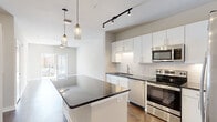Located at 128 Mill St in Clayton, NC, this one-bedroom, one-bathroom residence offers a well-designed layout across a single floor. The kitchen is a standout feature, showcasing white shaker cabinetry extending to the ceiling, a classic subway tile backsplash, dark granite countertops, a large center island with pendant lighting, and a full suite of stainless steel appliances including a side-by-side refrigerator, electric range, built-in microwave, and dishwasher. Track lighting adds a modern touch overhead. The open living area flows naturally from the kitchen and features hardwood-style flooring and neutral tones throughout, creating a bright and cohesive feel. A door leads directly to a private balcony, allowing natural light to fill the space. The bedroom is carpeted, generously proportioned, and includes a ceiling fan and a window with exterior views. The bathroom features white cabinetry, dark granite countertops, a framed mirror, a glass-enclosed shower, and access to a dedicated walk-in closet with built-in shelving. Soft, neutral wall tones and clean finishes are carried consistently throughout the entire residence.