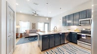 This single-story residence is designed with an open-concept floor plan that seamlessly connects the kitchen, dining, and living areas. The kitchen serves as a central focal point, outfitted with striking navy blue shaker-style cabinetry extending to the ceiling, crisp white quartz countertops, a subway tile backsplash, and a full suite of stainless steel appliances including a refrigerator, range, microwave, and dishwasher. A spacious island with seating for multiple bar stools provides additional prep space and casual dining. Pendant lighting illuminates the island, while recessed lighting and a ceiling fan enhance the open living area adjacent to the kitchen. Large windows dressed with sheer white curtains and wood blinds allow generous natural light to fill the space. A full-light glass entry door further contributes to the bright, airy atmosphere. The bedrooms are carpeted for comfort and feature large windows that invite natural light, while the bathroom connects directly to the main living corridor and showcases matching navy cabinetry with a wide vanity and white countertop. Light wood-look flooring flows throughout the main living areas, tying the design together with a cohesive, modern aesthetic.