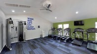 Located at 5600 Briar Oaks Ln in Raleigh, NC, this single-floor fitness center is designed to inspire and motivate residents toward an active lifestyle. The space features vaulted ceilings accented by recessed lighting and a ceiling fan, ensuring the room stays well-lit and comfortable during workouts. A bold motivational wall graphic — "Wake. Run. Lift. Eat. Sleep. Repeat." — sets an energizing tone against the crisp white walls, while a vibrant green accent wall adds a dynamic pop of color to the interior. The durable striped carpet flooring provides both cushioning and traction underfoot. The facility is equipped with a row of elliptical trainers and cardio machines positioned near natural-light-filled windows, as well as a multi-function strength training cable machine along the side wall. A wall-mounted television is also present for entertainment during workouts. Natural light filters in through double windows on the green accent wall, complementing the recessed overhead lighting to create a bright, open atmosphere. The overall design balances functionality and motivation, offering a well-equipped, inviting environment for residents to pursue their fitness goals year-round.