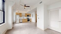 Located at 5000 Euclid Avenue in Cleveland's vibrant midtown corridor, this one-bedroom, one-bathroom unit offers a well-organized layout across a single floor. The open-concept living and kitchen area is anchored by a modern kitchen equipped with warm honey-toned wood cabinetry, stainless steel appliances including a refrigerator, dishwasher, microwave, and range, along with ample counter space and open shelving. Neutral-toned tile flooring runs throughout, lending a clean, cohesive aesthetic. A ceiling fan with integrated lighting adds both function and style to the main living space, which also benefits from track lighting overhead. The dedicated entrance opens into a hallway that leads to the spacious bedroom, which is generously sized and filled with natural light from a pair of large black-framed windows offering views of the Cleveland skyline and surrounding neighborhood. A closet provides convenient storage. The bathroom features a floor-to-ceiling white tile walk-in shower with brushed gold fixtures, offering a crisp, contemporary feel. In-unit laundry adds everyday convenience. The overall design embraces a minimalist, modern aesthetic with bright white walls and an airy atmosphere throughout.