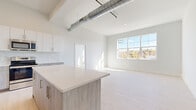This contemporary single-floor unit blends industrial character with modern finishes across a thoughtfully designed open layout. Soaring ceilings with exposed metallic ductwork lend an urban loft aesthetic to the expansive main living area, which is bathed in natural light from large multi-pane windows overlooking lush outdoor scenery. Light-toned hardwood flooring runs throughout, enhancing the bright, airy ambiance of every room.

The open-concept kitchen is a standout feature, anchored by a generous quartz-topped island with warm wood-toned cabinetry and gold hardware. Upper white cabinetry contrasts beautifully with a marble-effect tile backsplash, while stainless steel appliances — including a full-size refrigerator, range, and built-in microwave — complete the space. The kitchen flows seamlessly into the living area, making it ideal for both everyday living and entertaining.

The bedrooms are generously proportioned, each offering high ceilings, continued hardwood flooring, and large windows that invite natural light and outdoor views. The bathrooms are finished with striking marble-patterned tile on both the floors and tub surrounds, complemented by light wood vanities with brushed gold hardware, creating a clean, spa-like atmosphere. The overall design is cohesive, neutral, and refined throughout.