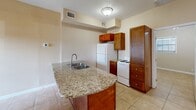 This efficient single-floor unit offers a well-organized layout comprising a kitchen, living area, one bedroom, and one bathroom. The kitchen serves as the focal point of the main living space, anchored by a curved granite-topped island with a double stainless steel sink and rich warm-toned wood cabinetry. A full-size refrigerator, electric range, and tall pantry cabinet provide practical storage and functionality. The kitchen flows openly into the living area, which is brightened by a glass-paned patio door that allows natural light in and is complemented by a ceiling fan overhead. Light beige walls and large-format ceramic tile flooring run consistently throughout the unit, creating a cohesive, clean aesthetic. The bedroom is a cozy, tile-floored room featuring a window with blinds that filters natural light, and a low built-in platform element along one wall. The bathroom includes matching warm-toned wood cabinetry with a granite vanity top, tile-surround shower, and overhead storage cabinet. HVAC vents are present throughout, ensuring climate control across the unit. Overall, the space presents a functional and consistent design with practical finishes suited for comfortable single-occupant living.