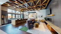 This thoughtfully designed single-floor amenity space offers a compelling blend of architectural character and versatile functionality. The centerpiece of the main lounge is a striking geometric coffered ceiling composed of warm wood beams set against a dark backdrop, lending visual depth and a refined industrial-meets-contemporary aesthetic. Large windows line the perimeter, flooding the open-plan interior with abundant natural light and offering views of the surrounding community.

The lounge area is furnished with eclectic, curated seating arrangements—including plush sofas, velvet accent chairs, and a bold area rug—centered around a dramatic feature wall clad in dark textured panels, which houses a wall-mounted television above a sleek linear gas fireplace. Pendant lighting adds warmth throughout.

A casual games area includes a full billiards table alongside a built-in bench with a textured upholstered backrest, complemented by high-top bar seating near the windows. A kitchenette with a light countertop, bar stools, and stainless steel appliances serves the space conveniently.

A separate conference room features a long communal wood table, upholstered chairs, a textured accent wall, curated artwork, and a modern pendant fixture, making it well-suited for meetings or collaborative gatherings.