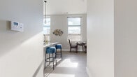This well-designed single-floor residence offers a thoughtfully laid-out living space with a cohesive modern aesthetic. The open-concept kitchen is a standout feature, boasting granite countertops, a large breakfast bar island, stainless steel appliances including a refrigerator, range, and microwave, and a mix of white upper cabinets and warm wood-toned lower cabinetry. Light-toned luxury vinyl plank flooring runs throughout the main living areas, reflecting an abundance of natural light that pours in through large windows. The kitchen flows seamlessly into the dining area, where a compact dining table sits beneath a pendant light, framed by views of the surrounding cityscape. Adjacent to the dining space, the living room offers a welcoming atmosphere accented by a bold blue and grey color palette, complete with a patterned area rug and modern furnishings. The bedroom is a calm retreat finished with plush carpet, clean white walls, and ample space for storage. The full bathroom features wood-look flooring, a soaking tub with shower, granite-topped vanity, and warm wood cabinetry, maintaining the consistent modern style found throughout the residence. Overall, the space strikes a balance between functionality and contemporary design.