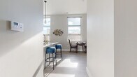 This well-designed single-floor residence offers a thoughtfully laid-out living space with a cohesive modern aesthetic. The open-concept kitchen is a standout feature, boasting granite countertops, a large breakfast bar island, stainless steel appliances including a refrigerator, range, and microwave, and a mix of white upper cabinets and warm wood-toned lower cabinetry. Light-toned luxury vinyl plank flooring runs throughout the main living areas, reflecting an abundance of natural light that pours in through large windows. The kitchen flows seamlessly into the dining area, where a compact dining table sits beneath a pendant light, framed by views of the surrounding cityscape. Adjacent to the dining space, the living room offers a welcoming atmosphere accented by a bold blue and grey color palette, complete with a patterned area rug and modern furnishings. The bedroom is a calm retreat finished with plush carpet, clean white walls, and ample space for storage. The full bathroom features wood-look flooring, a soaking tub with shower, granite-topped vanity, and warm wood cabinetry, maintaining the consistent modern style found throughout the residence. Overall, the space strikes a balance between functionality and contemporary design.