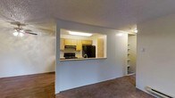 This single-story space is designed with an open-concept layout that promotes a seamless flow between the living and kitchen areas. The kitchen is partially enclosed and accessible via a pass-through opening in the dividing wall, offering both visual connectivity and practical counter space. It is equipped with light wood cabinetry, a black refrigerator, a black range with an overhead exhaust hood, and a single-basin sink, all illuminated by a recessed fluorescent ceiling light that brightens the workspace effectively. Built-in open shelving is visible adjacent to the kitchen, providing additional storage or display space. The living area features a combination of hardwood-style flooring near the kitchen transition and wall-to-wall carpet throughout the main space, adding warmth underfoot. A ceiling fan with an integrated light fixture is centrally mounted in the living area, contributing to both air circulation and ambient lighting. The walls are finished in a neutral light gray tone, complementing the textured popcorn ceiling. Overall, the space presents a functional and straightforward layout suited for everyday living.