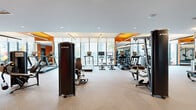 Located at 77 Christie St in Newark, NJ, this modern fitness center offers a generously sized, single-floor exercise space designed to support a comprehensive workout experience. The room is outfitted with a robust selection of professional-grade TRUE-branded cardio equipment, including treadmills, ellipticals, cycling bikes, and rowing machines, all arranged to maximize space and flow. A dedicated strength-training area features multiple cable and resistance machines, complemented by floor-to-ceiling mirrors that visually expand the room and allow users to monitor their form. The interior design is marked by bold orange accent walls and warm wood-toned ceiling panels that contrast with the clean white surfaces and neutral rubber flooring, creating an energizing yet polished atmosphere. Recessed and linear pendant lighting provides bright, even illumination throughout. A full wall of large-pane windows along one side floods the space with natural light while offering open views of the surrounding streetscape, enhancing the sense of airiness. Exposed brick detailing in the strength zone adds an industrial character that balances the contemporary finishes. The overall layout is thoughtfully organized to accommodate cardio, strength, and functional training zones within a single, well-appointed space.