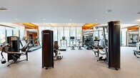Located at 77 Christie St in Newark, NJ, this modern fitness center offers a generously sized, single-floor exercise space designed to support a comprehensive workout experience. The room is outfitted with a robust selection of professional-grade TRUE-branded cardio equipment, including treadmills, ellipticals, cycling bikes, and rowing machines, all arranged to maximize space and flow. A dedicated strength-training area features multiple cable and resistance machines, complemented by floor-to-ceiling mirrors that visually expand the room and allow users to monitor their form. The interior design is marked by bold orange accent walls and warm wood-toned ceiling panels that contrast with the clean white surfaces and neutral rubber flooring, creating an energizing yet polished atmosphere. Recessed and linear pendant lighting provides bright, even illumination throughout. A full wall of large-pane windows along one side floods the space with natural light while offering open views of the surrounding streetscape, enhancing the sense of airiness. Exposed brick detailing in the strength zone adds an industrial character that balances the contemporary finishes. The overall layout is thoughtfully organized to accommodate cardio, strength, and functional training zones within a single, well-appointed space.