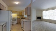 This single-floor space is designed with an open-concept layout that seamlessly connects the kitchen and living area, creating a cohesive and functional flow. The kitchen is equipped with light wood cabinetry, offering ample upper and lower storage, and features white appliances including an electric range and dishwasher. A double basin sink is positioned along the counter, which runs the length of the kitchen in a neutral tone that complements the cabinetry. An under-cabinet range hood is installed above the stove, and a pass-through window or opening connects the kitchen to the adjacent living space.

The living area transitions smoothly from tile flooring in the kitchen to plush carpeting, adding warmth underfoot. A tile-surround fireplace serves as a focal point in the living room, lending a cozy and inviting ambiance to the space. A window allows natural light to filter into the living area. The overall color palette is neutral throughout, with white walls and light-toned finishes creating a bright, clean aesthetic. The open design between the kitchen and living room enhances the sense of spaciousness across the single level.