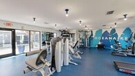 Located at The Oceanaire in Largo, Florida, this well-appointed fitness center offers a generously sized, single-floor space designed to meet a wide range of workout needs. The room is finished with durable blue rubberized flooring that provides both comfort and functionality, complementing the clean white walls and modern aesthetic throughout.

The cardio zone is equipped with multiple treadmills and an elliptical trainer, positioned along large floor-to-ceiling windows and glass double doors that invite ample natural light and provide refreshing views of the surrounding landscaping. A wall-mounted flat-screen television adds to the workout experience in this area.

The strength training section is comprehensively outfitted with a cable and pulley multi-station machine, a Hoist cable system, adjustable weight benches, and a full rack of dumbbells. An additional free weights area features a well-stocked rack of medicine balls and kettlebells, ideal for functional fitness training.

A striking branded mural in shades of blue and teal depicting tropical foliage and the "Oceanaire" name serves as a bold focal point on the rear wall, reinforcing the property's coastal identity. The space also includes a water fountain, a sanitizing station, and posted fitness center guidelines. Ceiling-mounted lighting ensures the entire area is well-lit at all hours.